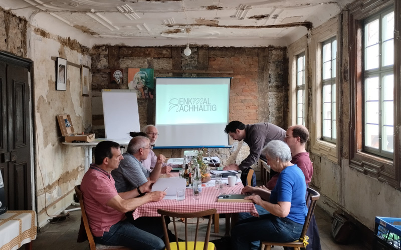 Sechs Personen sitzen an einem Tisch und arbeiten. Ein historisches Zimmer mit Stuck an der Decke. Im Hintergrund steht eine Leiwand.