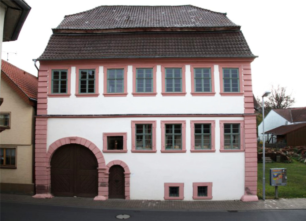 Historisches Fachwerkhaus in rosa und wei&szlig;er Farbe mit braunem Ziegeldach, sechs Fenstern und einem gro&szlig;en Toreingang, typisch f&uuml;r eine traditionelle deutsche Architektur in einem Dorf.