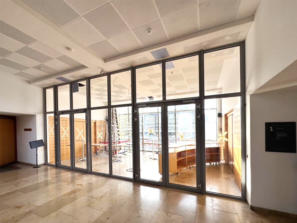 Gro&szlig;e Glasfront mit Fl&uuml;gelt&uuml;ren, die einen Eingangsbereich mit Marmorboden und modernem, offenem Interieur erschlie&szlig;en. Helle, luftige Architektur mit strukturierter Decke und seitlichen Wandelementen.