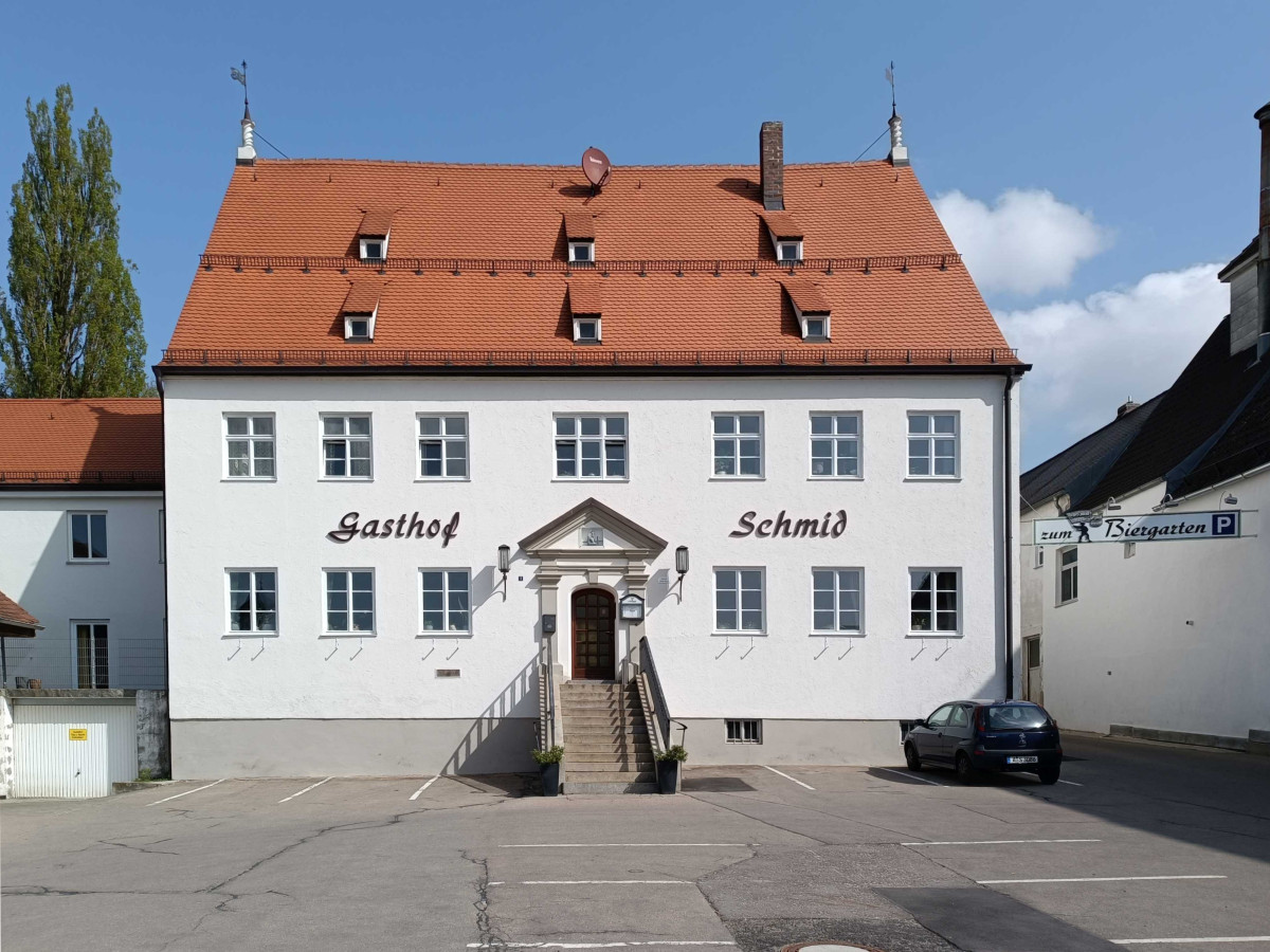 Wei&szlig;es zweist&ouml;ckiges Geb&auml;ude mit orangefarbenem Dach und der Aufschrift "Gasthof Schmid". Eine Treppe f&uuml;hrt zum Eingang, und vor dem Geb&auml;ude ist ein Parkplatz mit einem geparkten Auto.