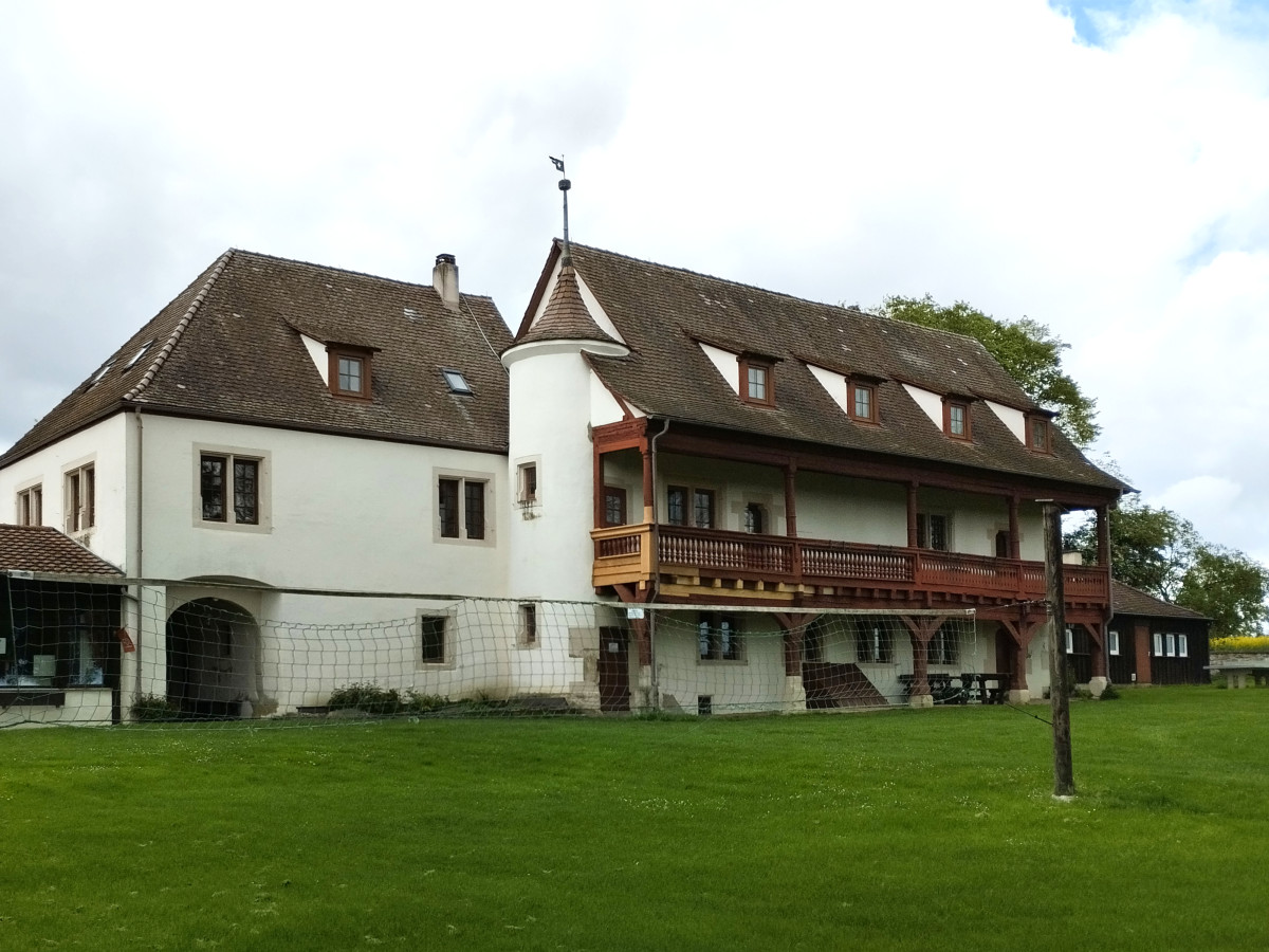 Historisches Hofgut Einsiedel mit wei&szlig;em Hauptgeb&auml;ude, umgeben von einer gr&uuml;nen Wiese und einigen B&auml;umen.