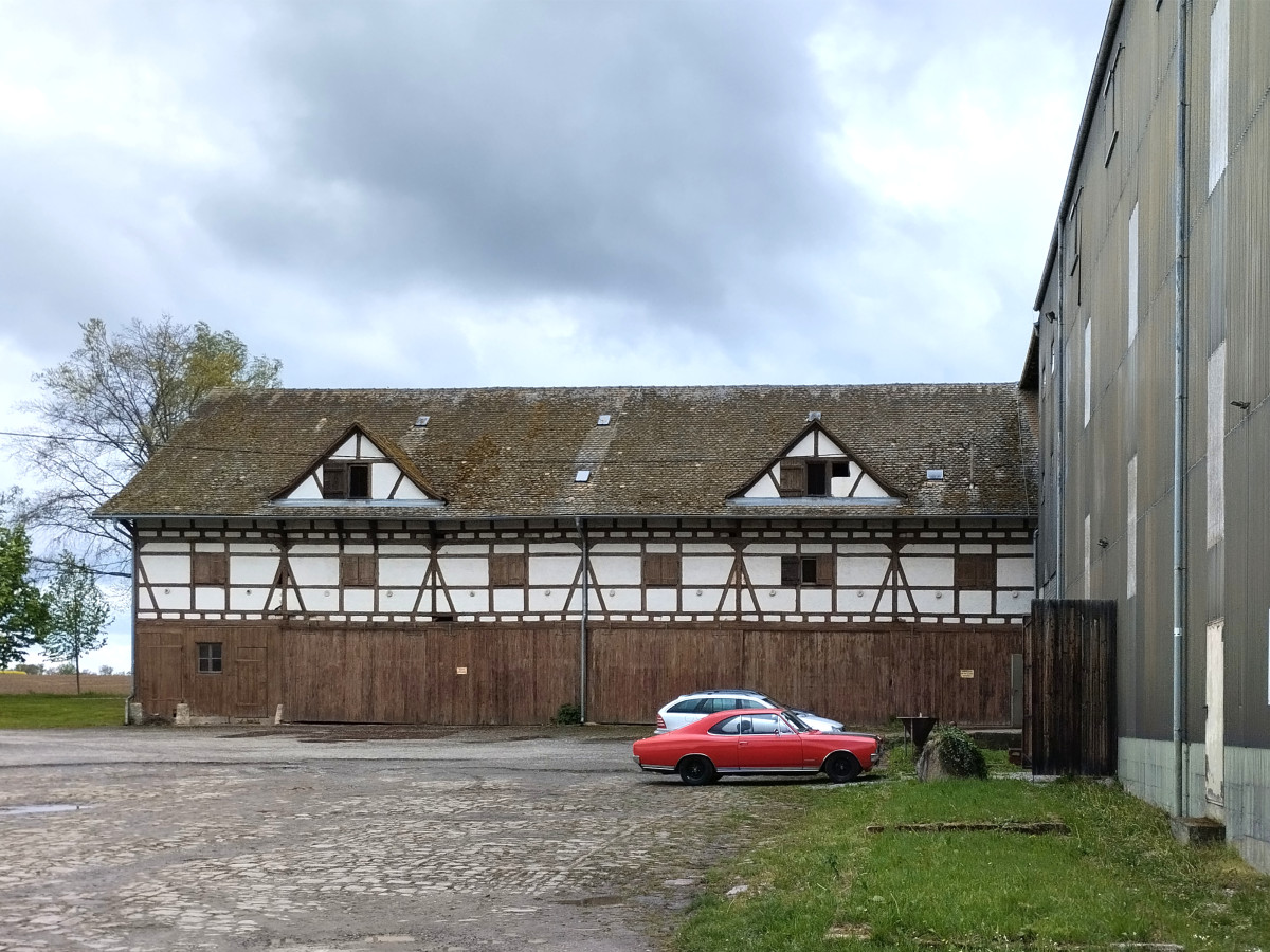 Historisches Fachwerkgeb&auml;ude mit rotem Fahrzeug davor, vor grauem Himmel auf einem l&auml;ndlichen Hof.