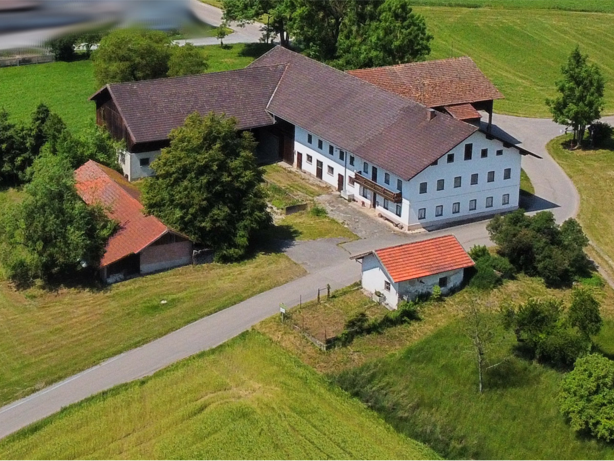 Luftaufnahme eines wei&szlig;en Bauernhofs mit mehreren Geb&auml;uden und roten D&auml;chern, umgeben von gr&uuml;nen Wiesen und B&auml;umen.