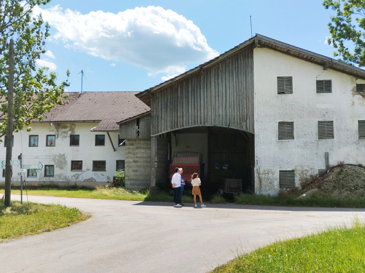 Zwei Personen stehen vor einem wei&szlig;en landwirtschaftlichen Geb&auml;udekomplex mit Holzscheune an einer asphaltierten Landstra&szlig;e, umgeben von gr&uuml;ner Wiese und B&auml;umen unter blauem Himmel mit wei&szlig;en Wolken.