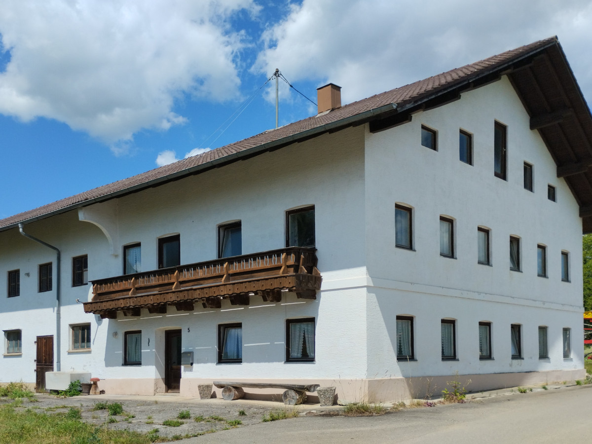 Zweist&ouml;ckiges wei&szlig;es Bauernhaus mit braunem Holzbalkon und Satteldach, umgeben von einer l&auml;ndlichen Umgebung mit blauem Himmel und Wolken.