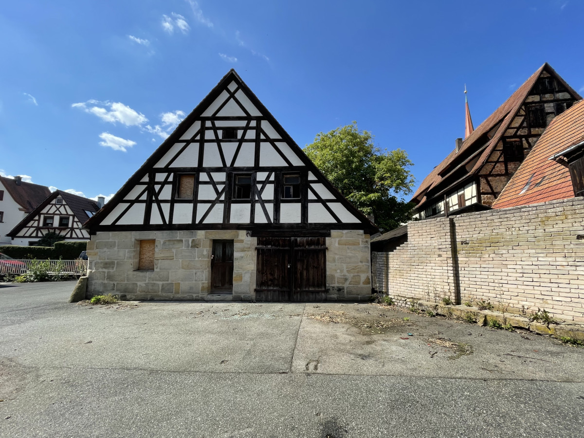 Historisches Fachwerkgeb&auml;ude mit schwarzen Holzbalken und Steinmauersockel auf einem Parkplatz, umgeben von weiteren traditionellen Fachwerkh&auml;usern unter blauem Himmel.