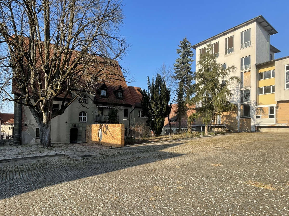 Blick auf das Kronenbr&auml;uareal mit historischen Geb&auml;uden und einem modernen Neubau, gepflasterter Stra&szlig;e und kahlen B&auml;umen vor blauem Himmel.