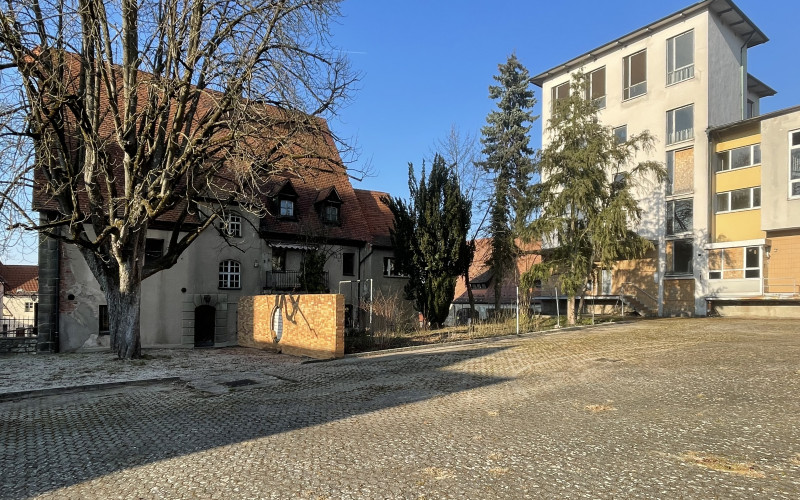 Blick auf das Kronenbräuareal mit historischen Gebäuden und einem modernen Neubau, gepflasterter Straße und kahlen Bäumen vor blauem Himmel.