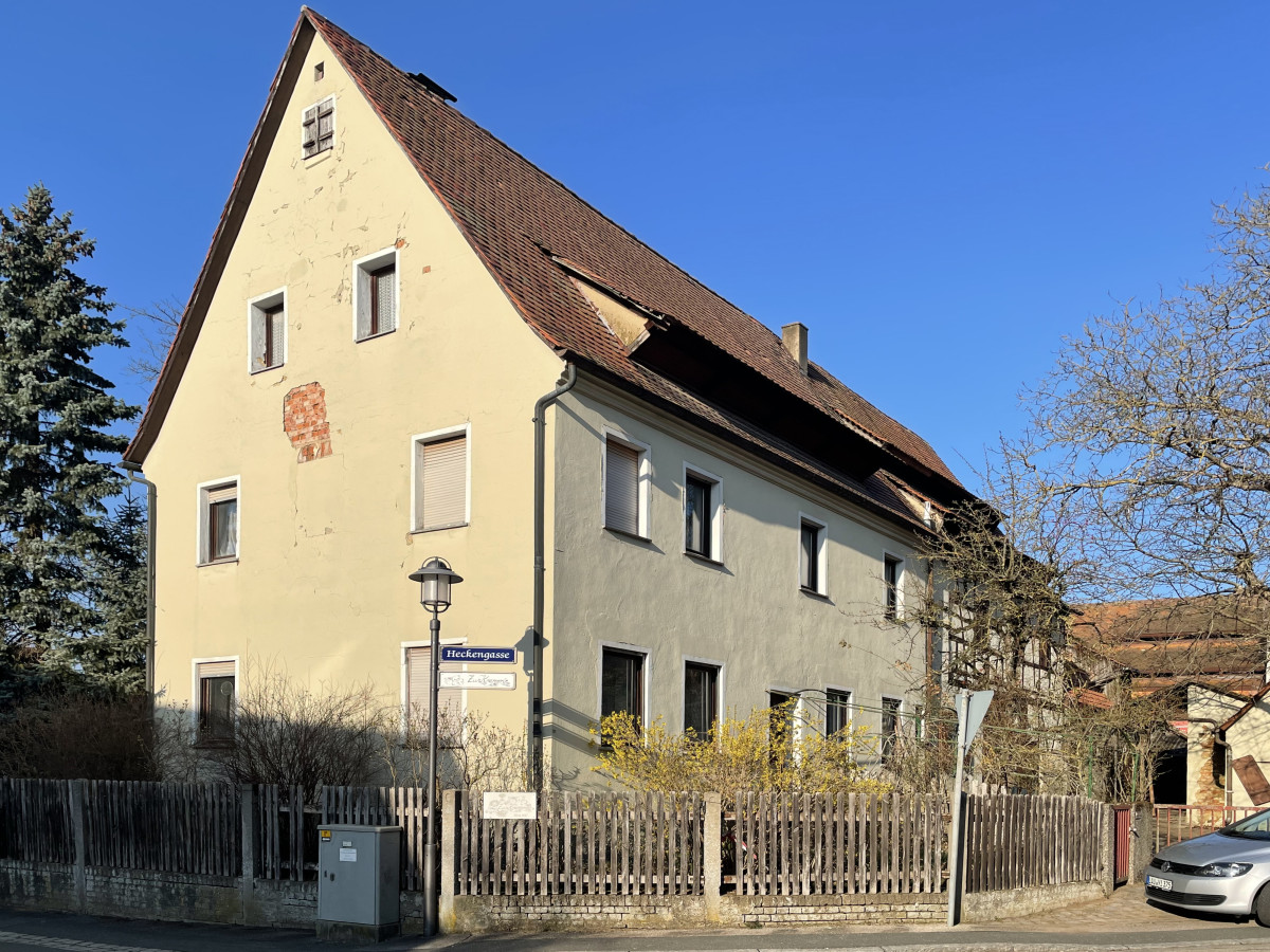 Gelbes Mehrfamilienhaus mit steilen Dach an einer Stra&szlig;e, umgeben von einem Holzzaun und B&auml;umen, vor blauem Himmel.