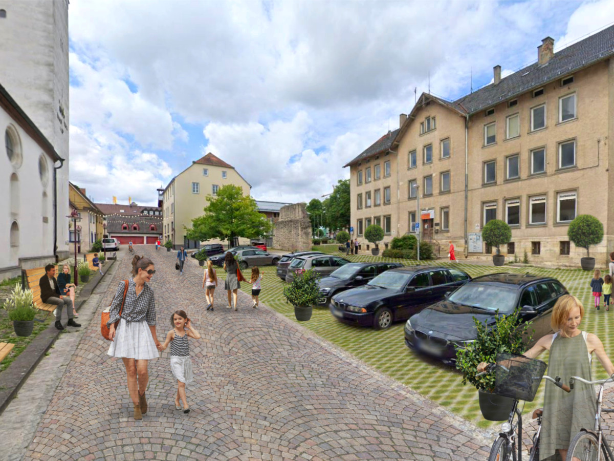 Kopfsteingepflasterter Stra&szlig;enbereich in einer kleinen Stadt mit Parkpl&auml;tzen, Wohnh&auml;usern und Fu&szlig;g&auml;ngern an einem bew&ouml;lkten Sommertag. Mehrere Autos sind entlang einer begr&uuml;nten Fl&auml;che geparkt, w&auml;hrend Menschen den Gehweg entlang gehen.