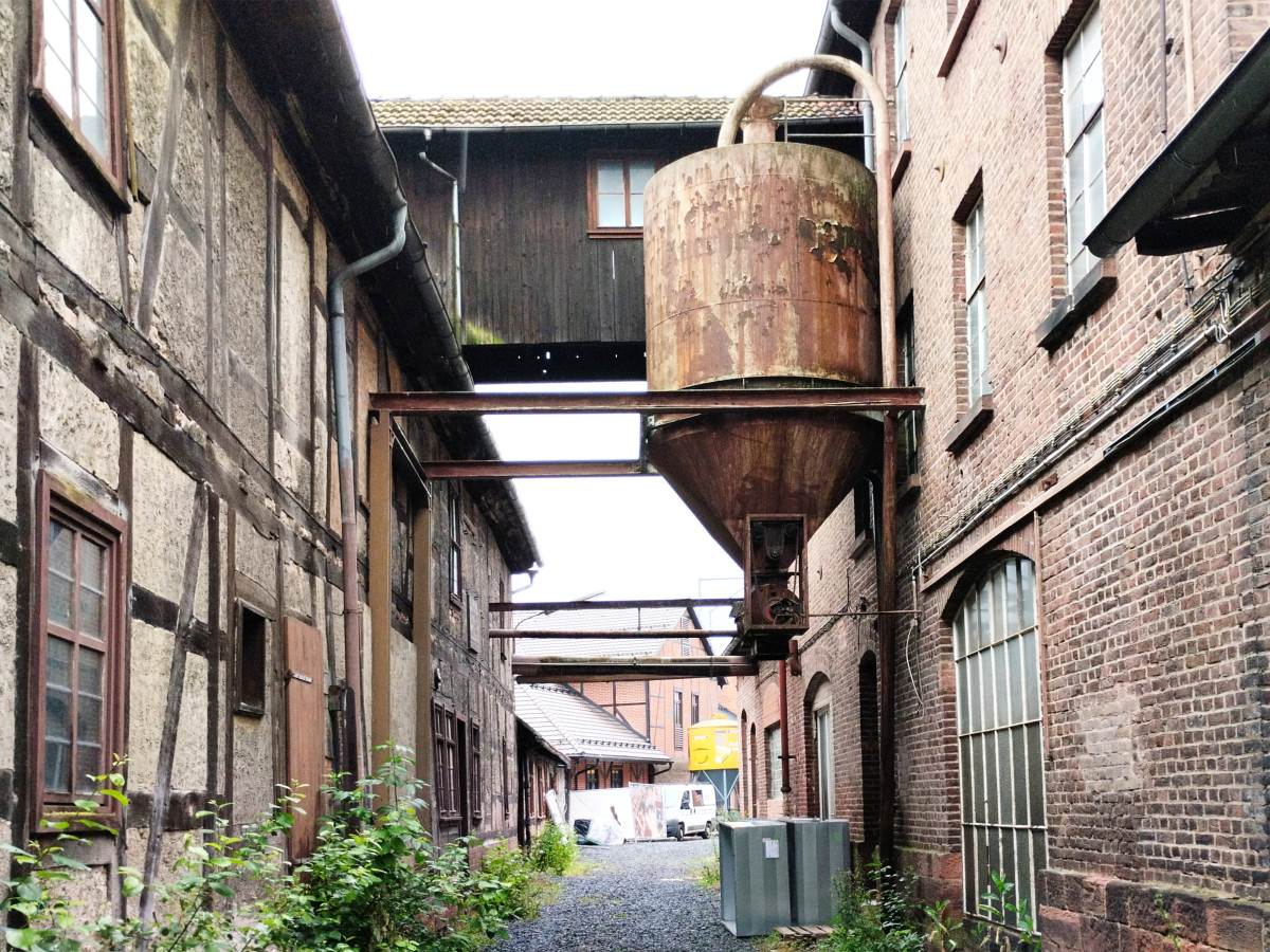 Alter Industriehof mit rostigem Wassertank, verbunden durch Stahltr&auml;gerbr&uuml;cken zwischen ziegligen und verwitterten Geb&auml;uden. Enge Gasse mit Unkraut und einem Container im Vordergrund, die eine industrielle Vergangenheit erahnen l&auml;sst.