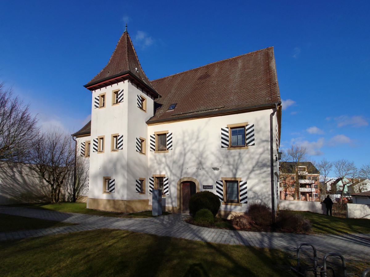 Wei&szlig; verputztes Schl&ouml;ssle mit schwarzem Satteldach und markanter Eckturm, umgeben von kahlen B&auml;umen vor blauem Himmel. Die Fenster sind mit schwarz-wei&szlig;en Fensterl&auml;den geschm&uuml;ckt, die dem Geb&auml;ude eine historische Ausstrahlung verleihen.
