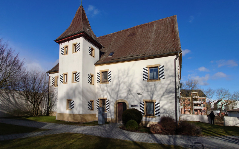 Weiß verputztes Schlössle mit schwarzem Satteldach und markanter Eckturm, umgeben von kahlen Bäumen vor blauem Himmel. Die Fenster sind mit schwarz-weißen Fensterläden geschmückt, die dem Gebäude eine historische Ausstrahlung verleihen.