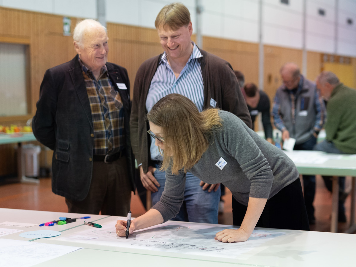 Teilnehmerin skizziert etwas auf einem gro&szlig;en Dokument, w&auml;hrend zwei &auml;ltere Herren im Hintergrund zusehen. Sie scheinen an einem Workshop oder einer Planungssitzung teilzunehmen.