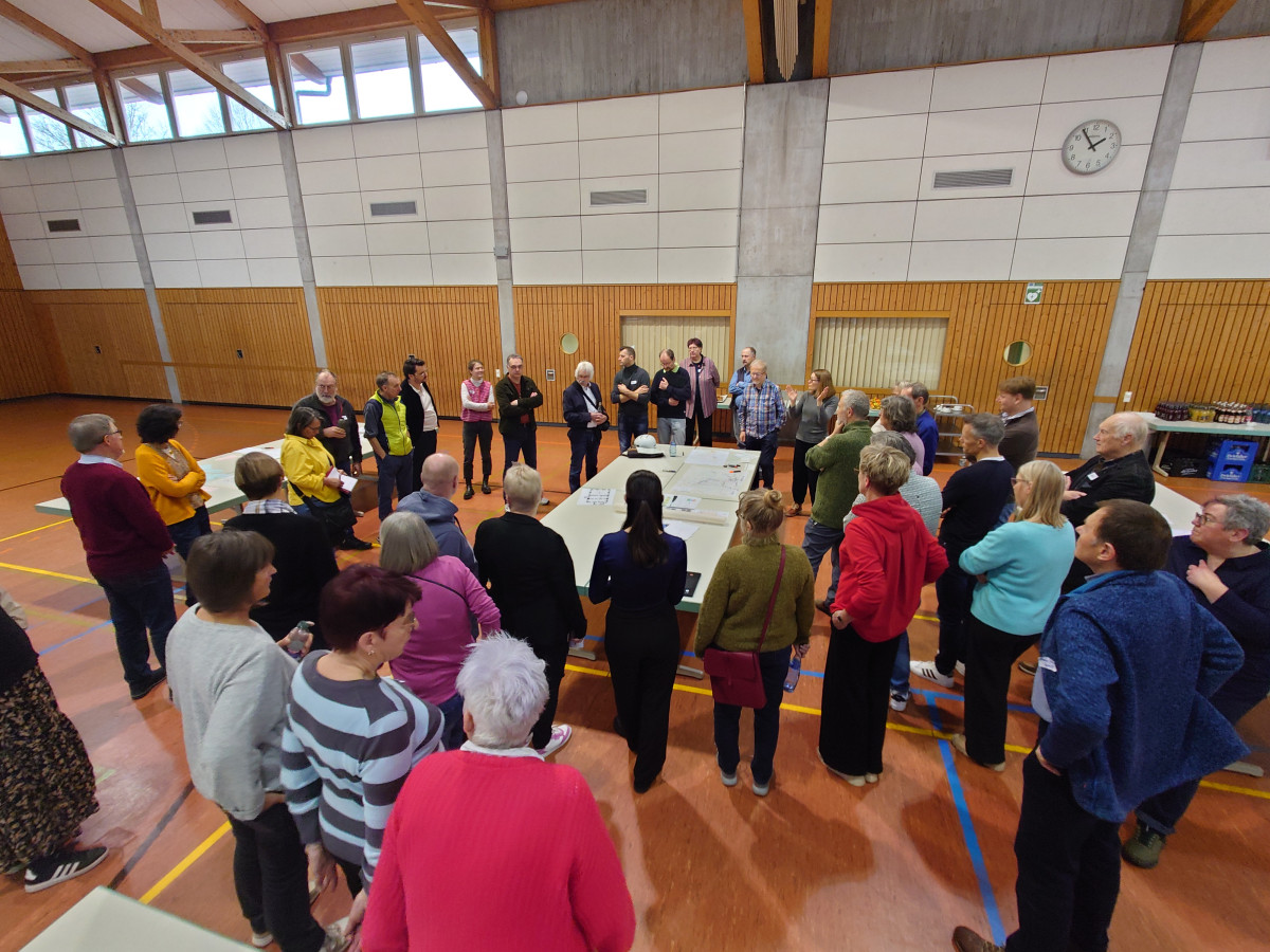 Gro&szlig;e Gruppe von Menschen in einer Sporthalle, die in einem Halbkreis um einen zentralen Tisch stehen und einer Pr&auml;sentation oder Besprechung beiwohnen.