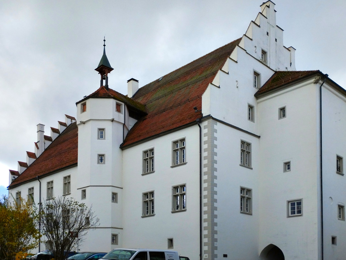Wei&szlig;es historisches Geb&auml;ude mit rotem Ziegeldach, Treppengiebeln und einem runden Turm, typisch f&uuml;r eine mittelalterliche Stadtarchitektur. Das Geb&auml;ude hat mehrere Fenster und steht in einer st&auml;dtischen Umgebung mit einem parkenden Fahrzeug im Vordergrund.