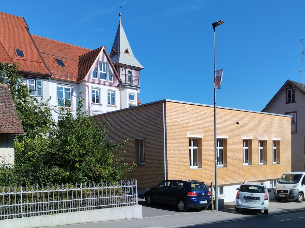 Historisches Geb&auml;udeensemble mit einem &auml;lteren Fachwerkhaus mit Turm und einem modernen Backsteingeb&auml;ude, davor parkende Autos an einem sonnigen Tag mit blauem Himmel.