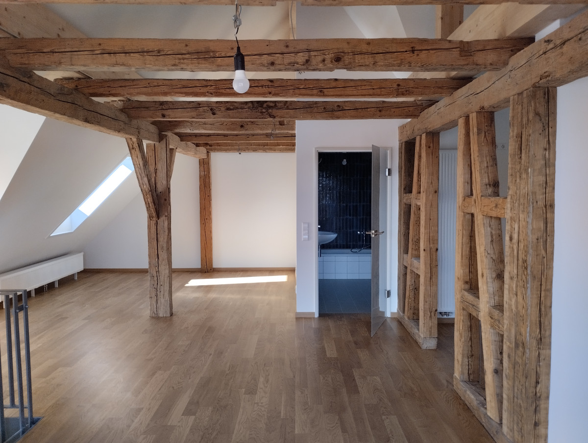 Dachgeschossraum mit Holzbalkendecke und -w&auml;nden, heller Holzboden, wei&szlig;e W&auml;nde und ein Fenster mit Tageslicht. Blick auf eine blaue T&uuml;r, die zu einem angrenzenden Raum f&uuml;hrt.