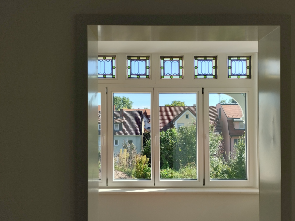 Modernes, mehrteiliges Fenster mit Sprossenfenstern im Oberbereich, Blick auf H&auml;user und G&auml;rten im Au&szlig;enbereich. Die Fensterrahmen sind wei&szlig;, und die Scheiben spiegeln das Tageslicht wider.