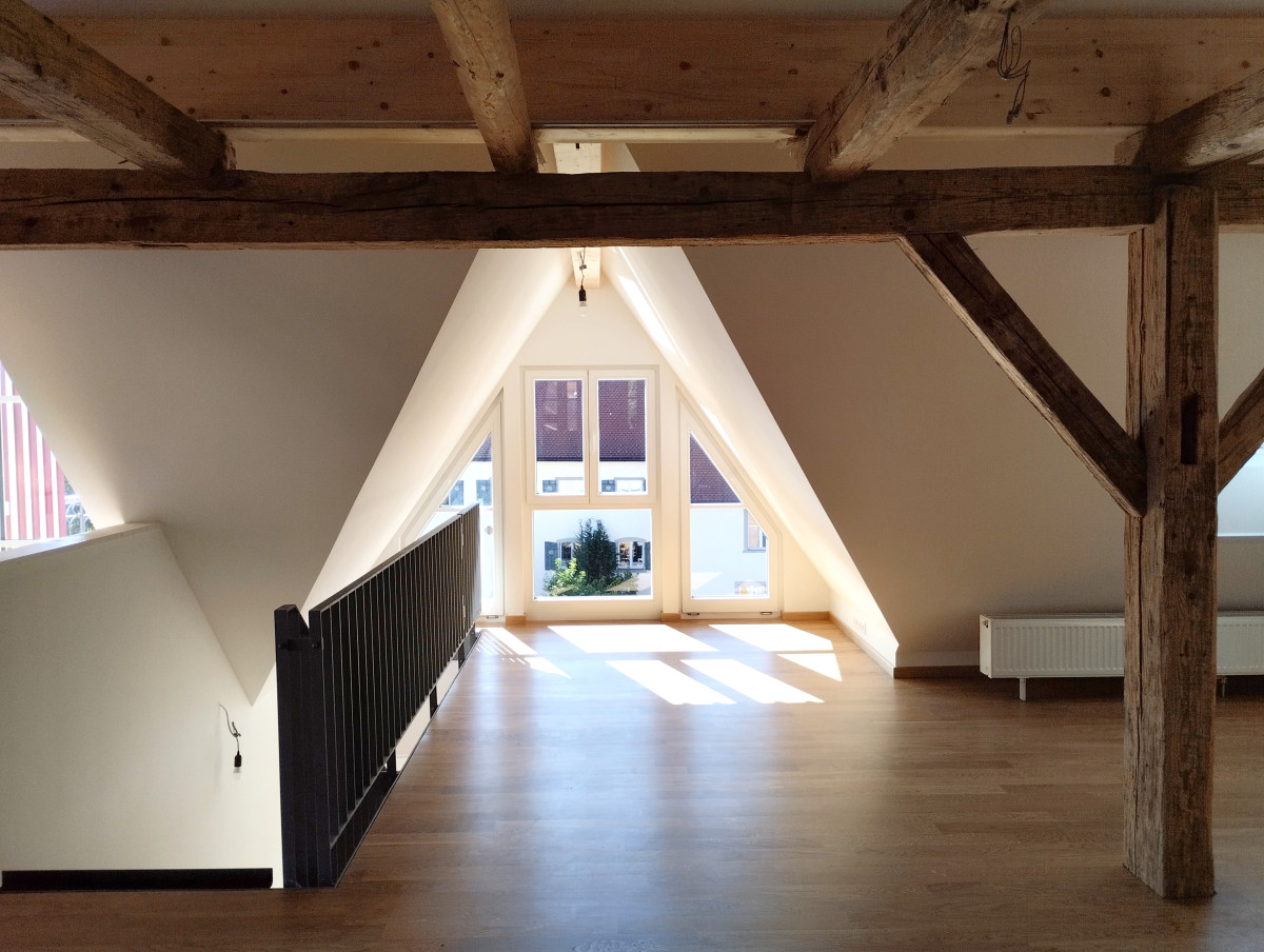 Dachgeschossraum mit Holzbalken und gro&szlig;em Dreiecksfenster, hell erleuchtet mit Holzboden und minimalistischer Einrichtung. Schwarzes Gel&auml;nder links und wei&szlig;e Sitzgelegenheit rechts, Sonnenlicht f&auml;llt durch das Fenster ein.