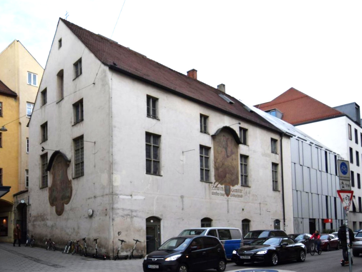 Altes dreist&ouml;ckiges wei&szlig;es Steingeb&auml;ude an einer Stra&szlig;enecke mit mehreren Fensterebenen und angrenzenden modernen Geb&auml;uden. Schwarze Autos parken entlang der Stra&szlig;e vor dem historischen Haus.