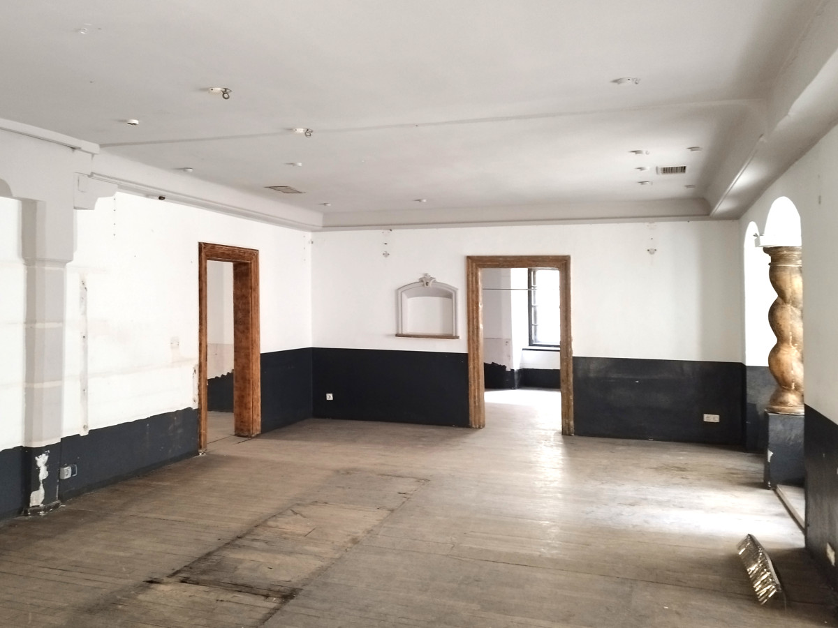 Leerer Raum mit wei&szlig;en W&auml;nden und schwarzem Sockel, zwei Holzt&uuml;ren und einer Bodenskulptur an der Seite. Der Raum hat einen unebenen Betonboden und Deckenspots.